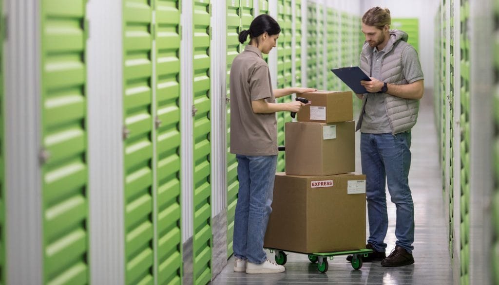 Two workers with stacked cardboard boxes on a dolly inside a storage facility with green doors.