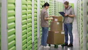 Two workers with stacked cardboard boxes on a dolly inside a storage facility with green doors.