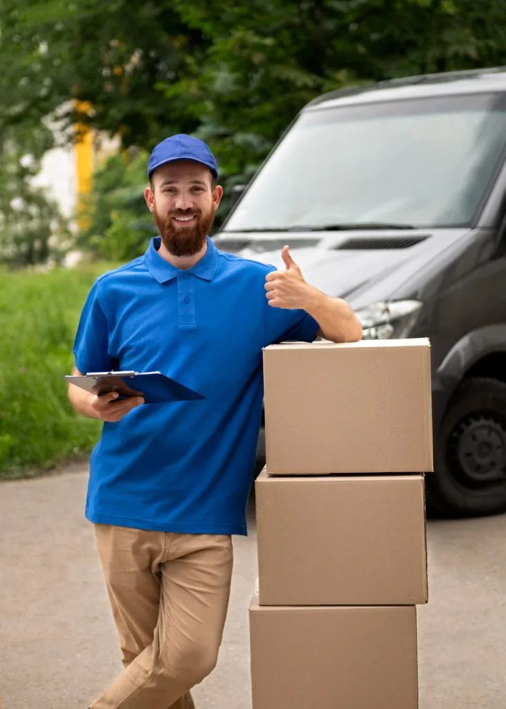 Smiling delivery man in blue uniform giving thumbs up next to stacked cardboard boxes and a van.