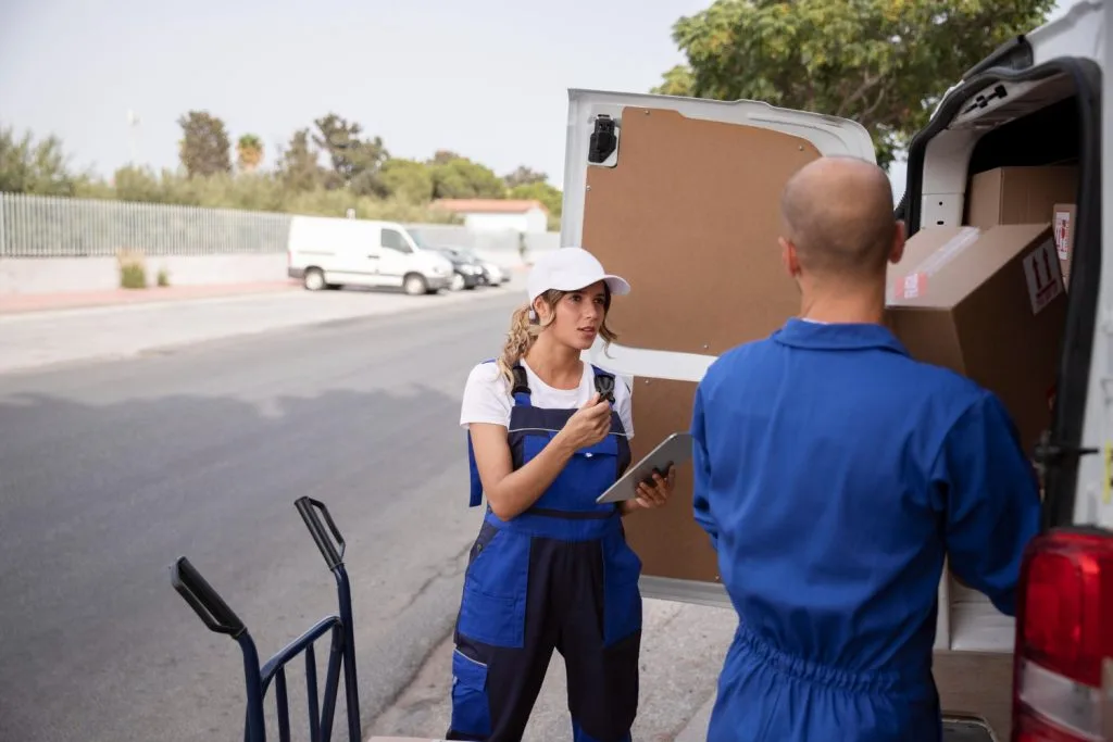Two movers in blue uniforms working beside a van, one holding a clipboard and talking while the other loads boxes.